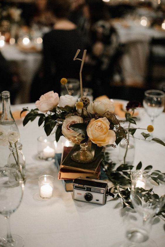 a stylish eclectic wedding centerpiece of a book stack, a vintage camera, candles and a pastel and neutral bloom arrangement with a table number