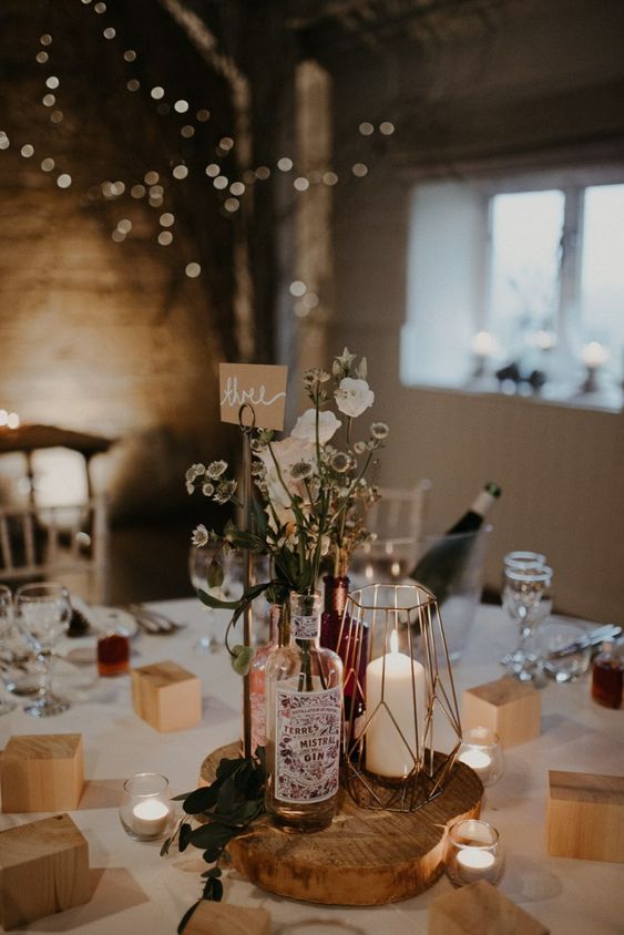 a whimsy wedding centerpiece of a wood slice, bottles with greenery and white blooms, a copper candle lantern and a table number