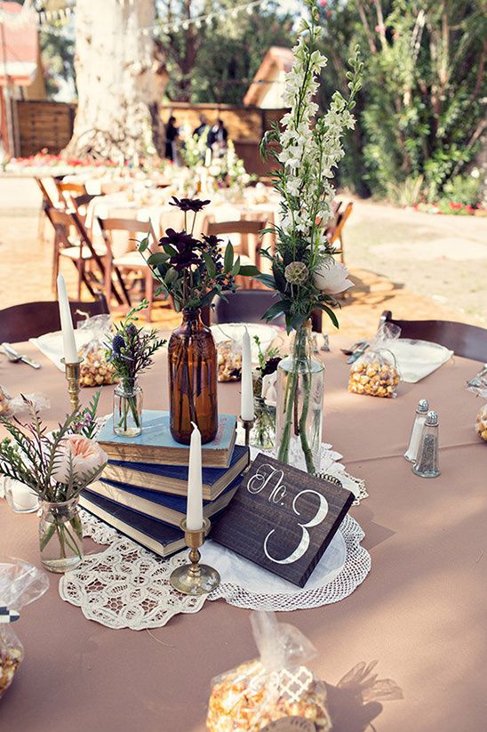 an eclectic boho wedding centerpiece of a doily, a book stack, bottles with bold blooms and greenery and a candle plus a table number