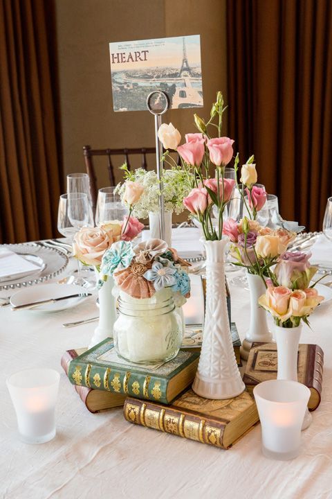 an eclectic vintage wedding centerpiece of book stacks, white vases with pastel blooms and a jar with pastel fabric flowers