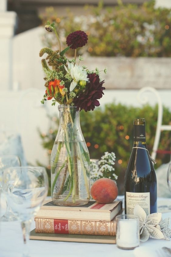 an eclectic wedding centerpiece of a book stack, a peach, candles, a bold floral arrangement in a bottle