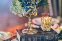 an eclectic wedding centerpiece of a book stack, gilded candleholders, a vase with bold blooms and greenery
