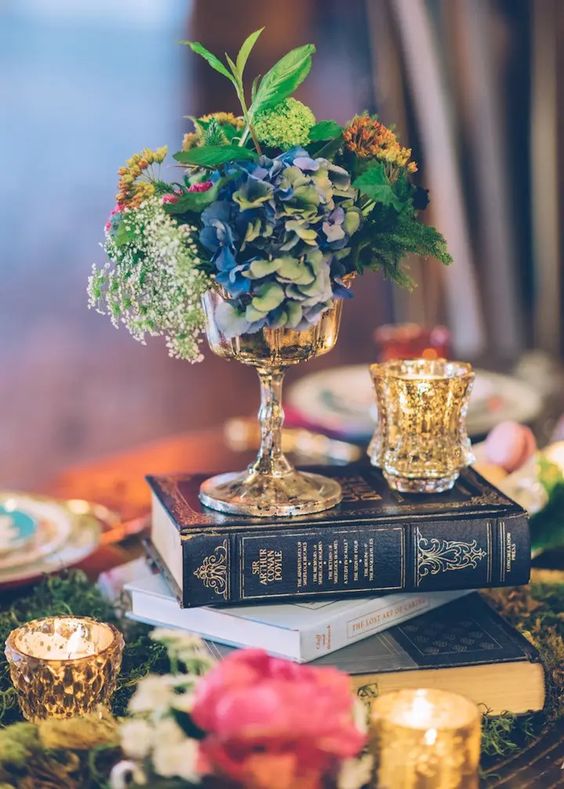 an eclectic wedding centerpiece of a book stack, gilded candleholders, a vase with bold blooms and greenery