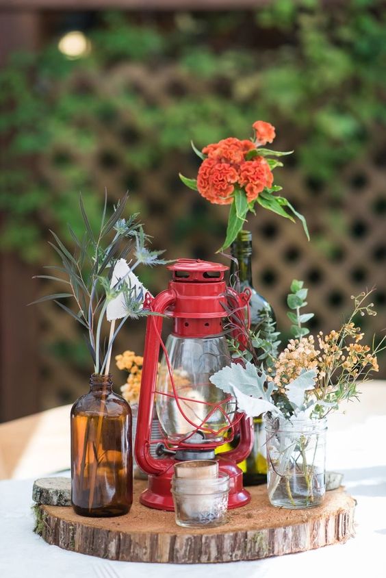 an eclectic wedding centerpiece of a tree slice, candleholders and a hurricane lantern, bottles and jars with mismatching blooms