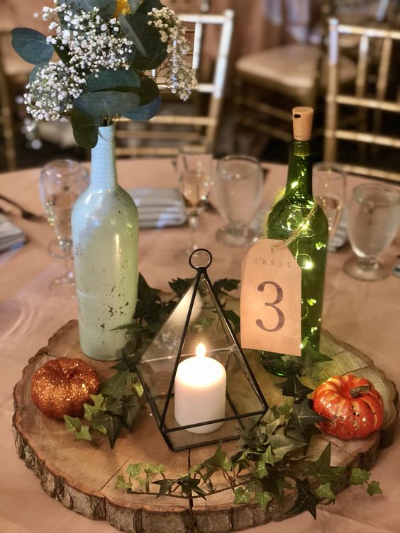an eclectic wedding centerpiece of a tree slice with greenery, glitter pumpkins, a terrarium candleholder, a bottle with blooms and leaves and a bottle with lights