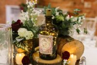 an eclectic wedding centerpiece of tree slices, candles, a bottle with LED lights, some neutral and burgundy blooms for a rustic wedding