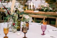 an eclectic wedding centerpiece of wine and beer bottles, greenery, neutral and pastel blooms and grasses is great for a boho wedding