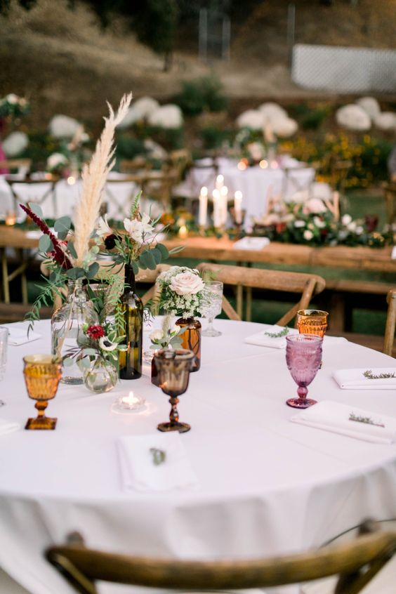 an eclectic wedding centerpiece of wine and beer bottles, greenery, neutral and pastel blooms and grasses is great for a boho wedding