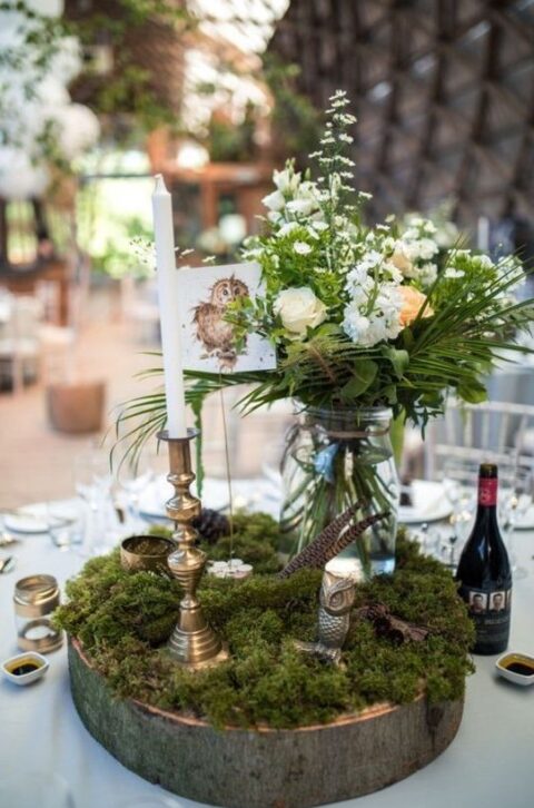 an enchanted forest wedding table setting with a wood slice, moss, a tall and thin candle, a jar with greenery and neutral blooms and feathers