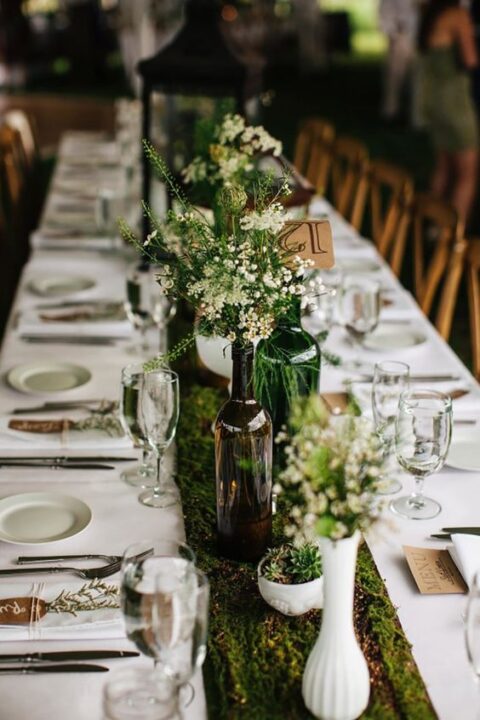 an enchanted forest wedding tablescape done with white linens, a moss table runner, blooms and potted succulents, a candle lanter is a lovely idea