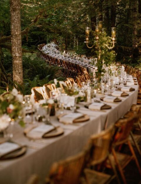 an extra long curved enchanted forest wedding table with tall candelabras covered with greenery and some blooms and wood slices as placemats
