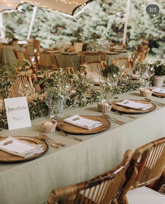 a cozy rustic wedding tablescape with a sage green tablecloth, greenery, baskets with bread, wooden placemats and chic cutlery