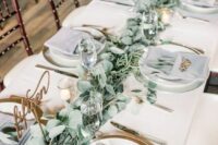 22 a delicate wedding tablescape with a sage green table runner, grey napkins and greenery, gold cutlery and calligraphy