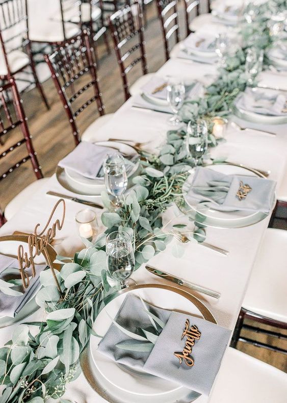a delicate wedding tablescape with a sage green table runner, grey napkins and greenery, gold cutlery and calligraphy