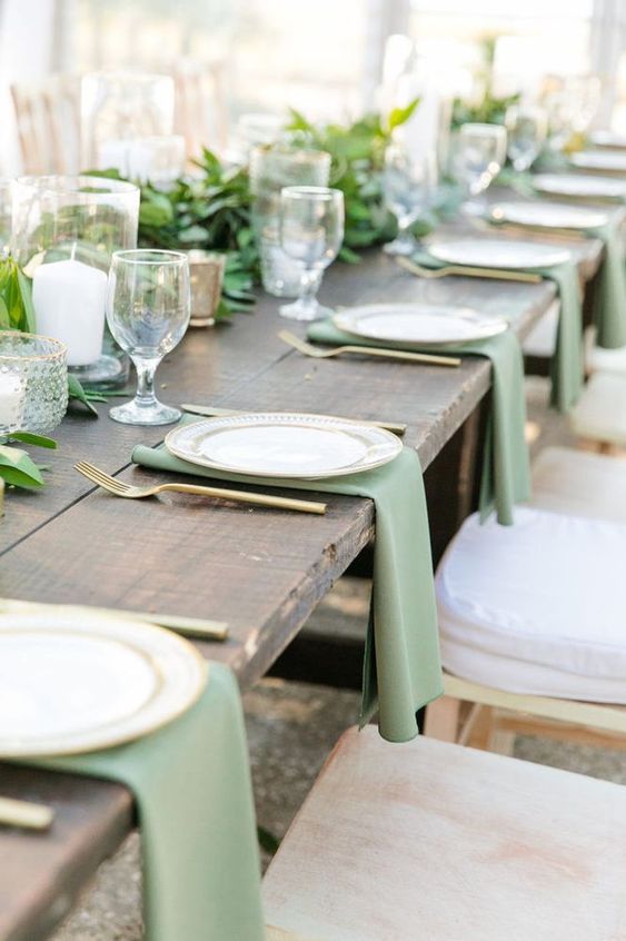 a lovely rustic wedding tablescape with an uncovered table, sage green napkins and greenery, pilalr candles and gold cutlery
