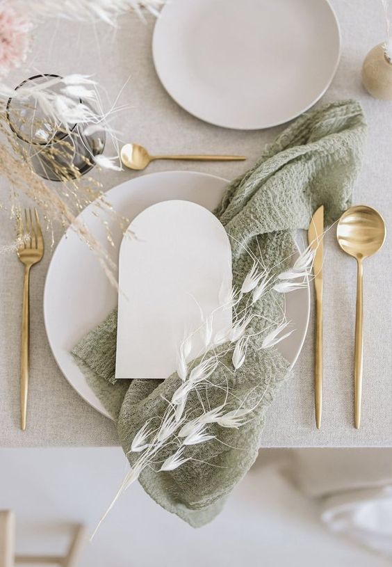 a modern rustic wedding tablescape with a neutral tablecloth, neutral porcelain, a sage green napkin and some dried herbs