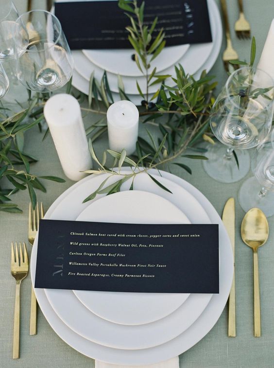 a refined modern wedding table setting with a sage green tablecloth, white porcelain, a black menu, gold cutlery and greenery plus pillar candles