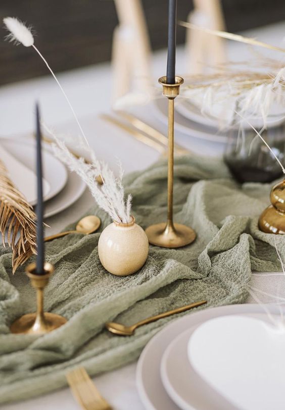 a rustic boho wedding tablescape with a sage green table runner, black candles in gold candleholders, gold cutlery and neutral porcelain