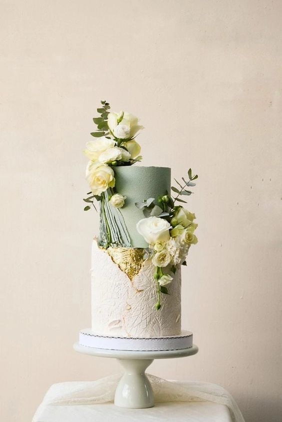 a catchy wedding cake with a textured white tier and a sage green one, gold leaf, white blooms and greenery