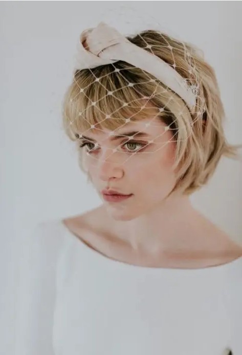 a short blonde bob with bangs, a knot headband and a birdcage veil for a retro touch to the look