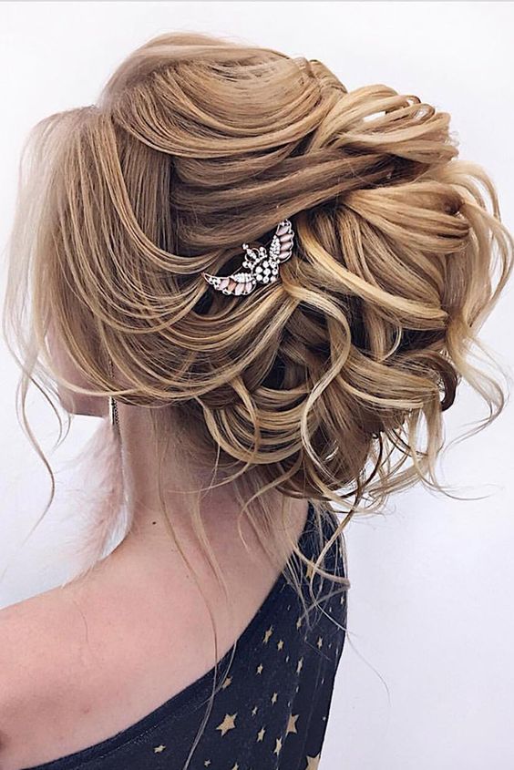 a sophisticated wavy wedding updo with a lot of hair, a volume on top and some locks down plus a rhinestone hairpiece
