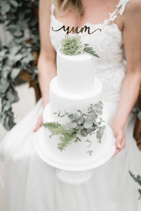 a white wedding cake decorated with sage green elements and a gold calligraphy topper