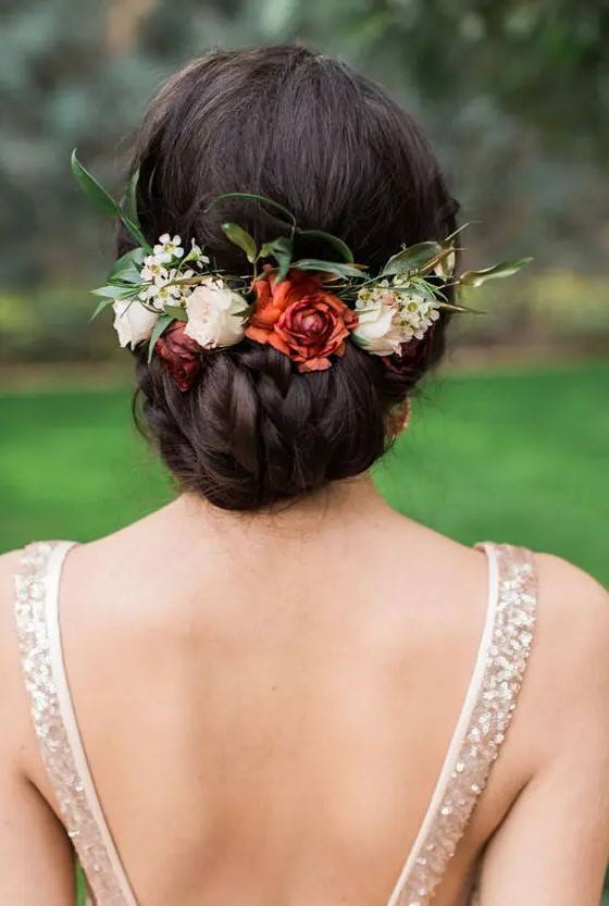 a braided and twisted low updo with a volume on top, white and rust blooms and greenery tucked in is a great idea for a fall bride