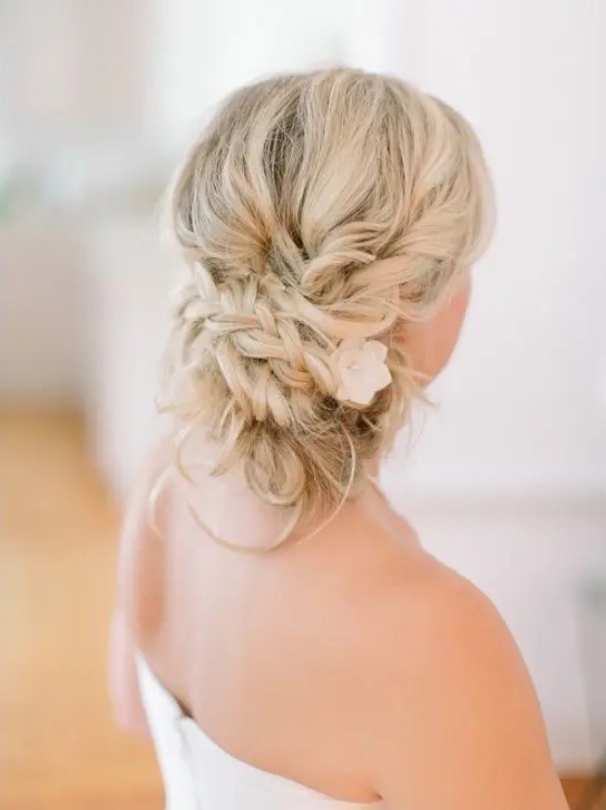 a braided messy low updo with a flower tucked inside and some little curls is a relaxed and chic solution for a boho bride