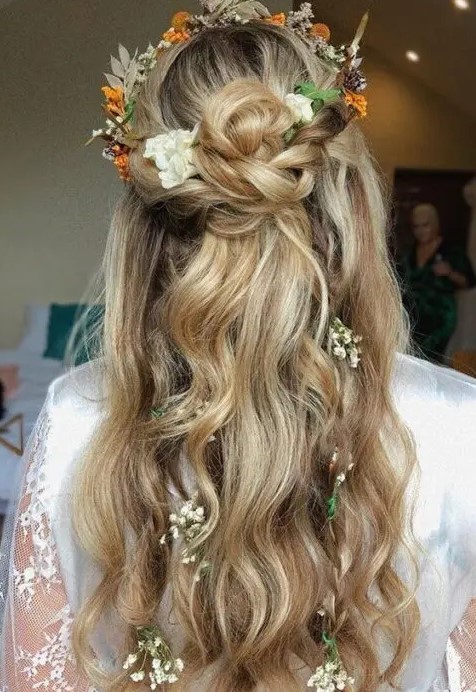 a fall boho wedding half updo with a twisted braided halo and a top knot, with waves down, a dried flower crown and more blooms in the hair
