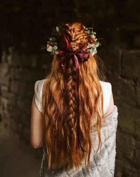 a pretty half updo with messy and textural braids on top and wavy locks down plus a greenery and fall floral crown with a burgundy bow