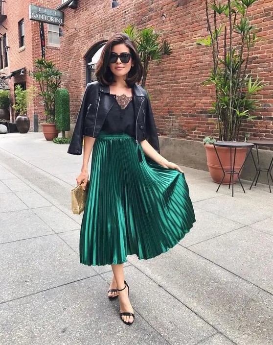 a black top with a lace neckline, a black cropped leather jacket, a green metallic pleated midi skirt, black heels