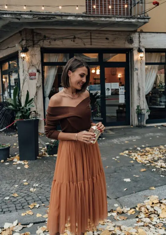 An elegant fall wedding guest outfit with a brown off the shoulder top, a rust colored tulle midi skirt and a small metallic clutch