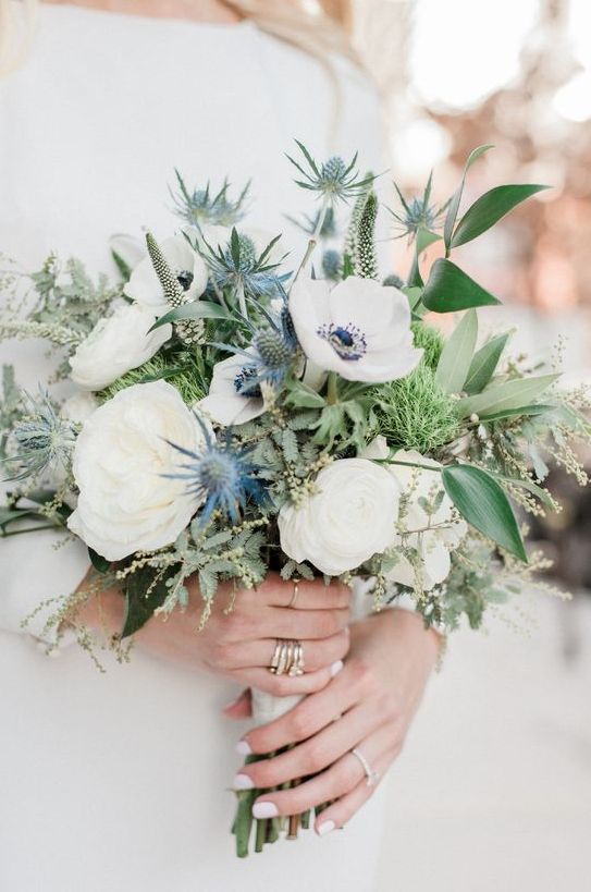 a delicate wedding bouquet of white roses and anemones, blue thistles and various types of greenery for a spring or summer wedding