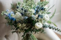 29 a dimensional and textural wedding bouquet of white ranunculus, waxflower, fillers, blue blooms, thistles and greenery