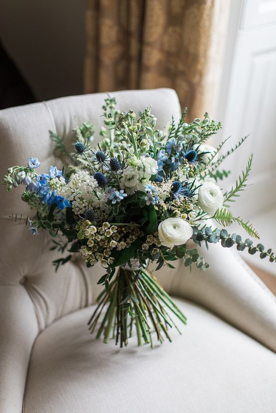 a dimensional and textural wedding bouquet of white ranunculus, waxflower, fillers, blue blooms, thistles and greenery