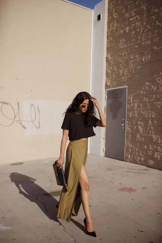 A modern and casual wedding guest look with a black crop top, a mustard colored maxi skirt with a thigh high slit, black shoes and a clutch