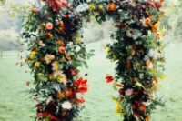a beautiful and colorful rustic fall wedding arch with greenery, red, yellow, pink blooms, including dahlias and mums, colorful fall leaves is a lovely idea for the season