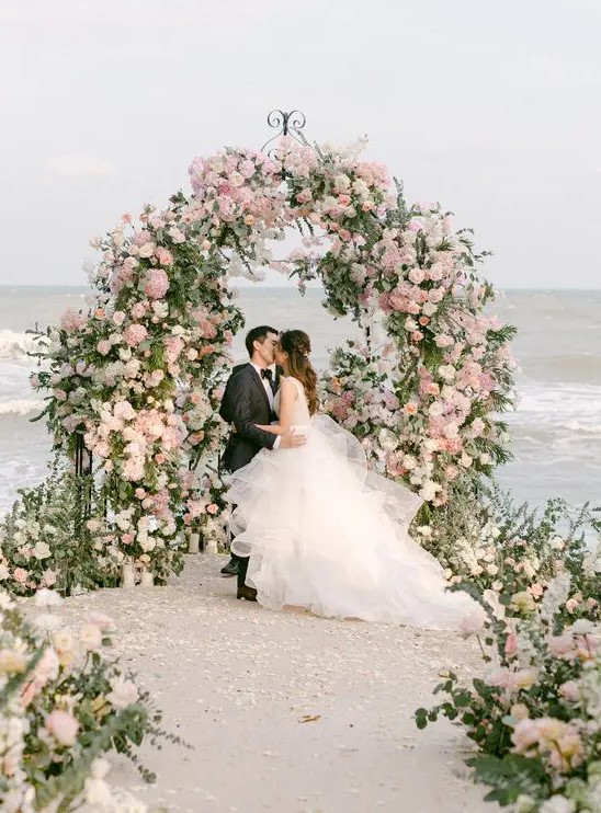 a beautiful and lush curved wedding arch covered with greenery, neutral and blush blooms and with a matching wedding aisle plus a sea view