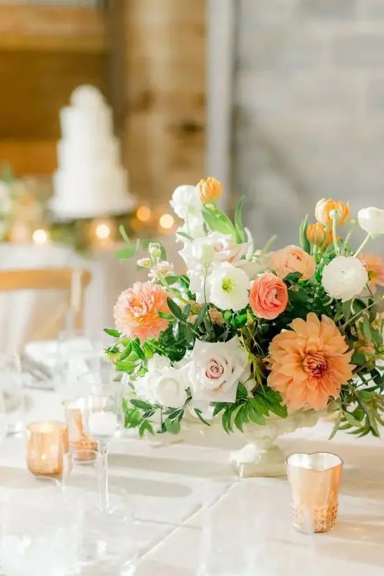 a beautiful earthy wedding centerpiece of white and orange ranunculus, blush and white roses and orange dahlias plus greenery for a summer or fall wedding