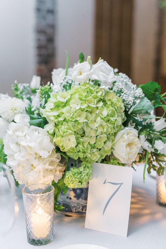 a beautiful wedding centerpiece of green and white hydrangeas, white roses and some fillers plus candles around