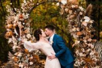 a boho lux fall wedding arch decorated with blush, rust-colored, peachy blooms, fronds, grasses, leaves and a baby’s breath