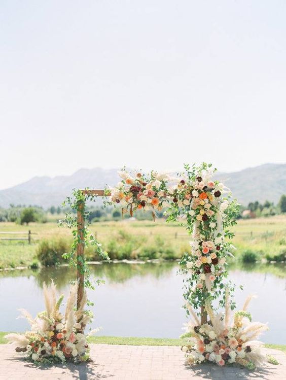 a boho wedding arch done with greenery, white, orange, pink and burgundy dahlias and some pampas grass plus a river view