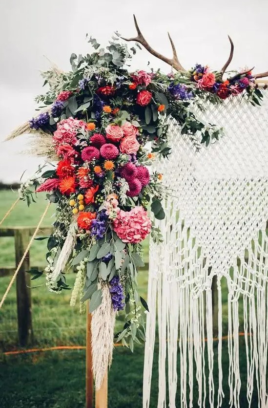 a boho wedding arch with a macrame hanging, antlers, pink, orange, lilac and purple blooms, greenery and grasses