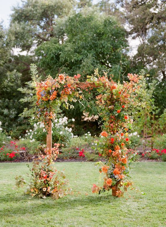 a bold fall wedding arch decorated with greenery, orange and red dahlias, twigs and branches is a stylish idea to rock