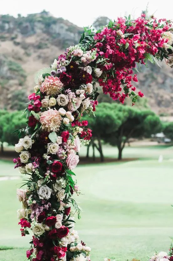 a bold jewel tone wedding arch with deep red, burgundy and fuchsia blooms, white and blush ones plus some greenery