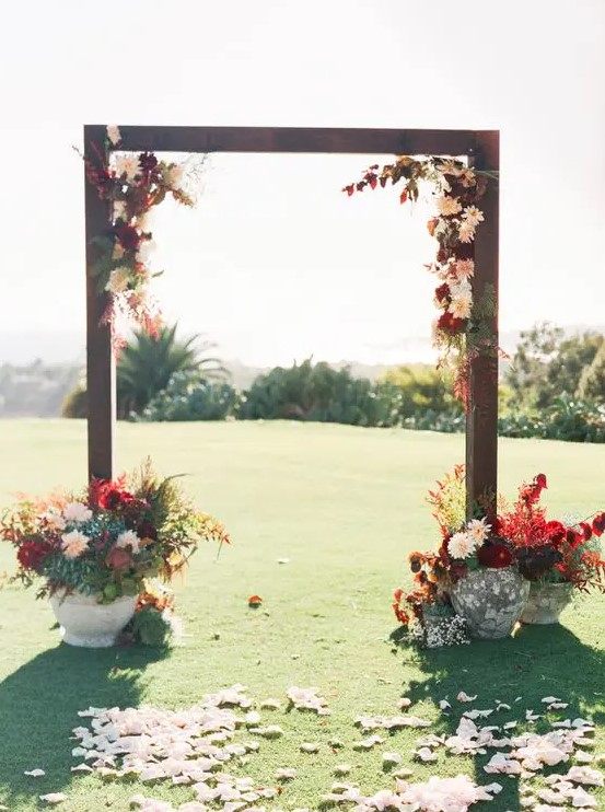 a bold rustic fall wedding arch decorated with burgundy and blush blooms, greenery and bold foliage, with matching arrangements in vases at the base
