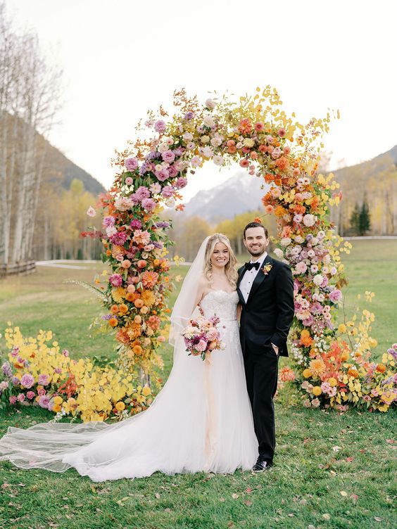 a bold wedding arch with orange, rust, yellow, pink and blush flowers and fall leaves and dahlias and roses is a lovely idea