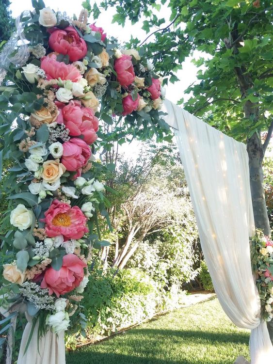 a bold wedding arch with sheer fabric, coral peonies, white and blush roses and greenery is a stylish idea for a wedding