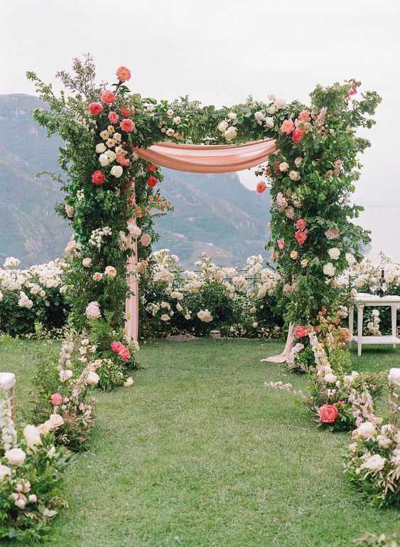 a bright and chic wedding arch done with greenery, white and blush blooms and coral peonies is a lovely idea, and a mountain view completes the look