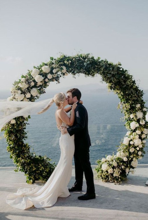 a chic round wedding arch with greenery and white blooms on some parts of it looks stylish and out of the box